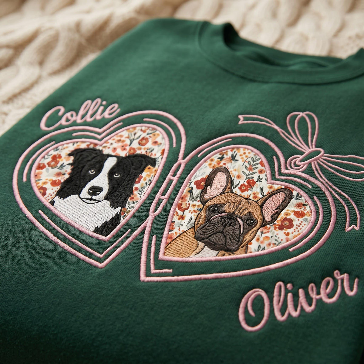 A dark green sweatshirt featuring an embroidered pink locket design with two heart-shaped frames. Inside the frames are floral patterns and portraits of a black and white dog and a fawn French Bulldog. The names Collie and Oliver are stitched.
