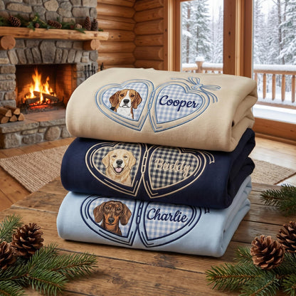 A stack of three folded sweatshirts in beige, navy blue, and light blue. Each features an embroidered heart-shaped locket design with a dog portrait on the left and a name on the right: Cooper, Buddy, and Charlie in script.