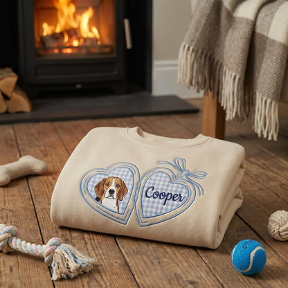 A beige crewneck sweatshirt featuring an embroidered design of an open heart-shaped locket. The left side of the locket displays a beagle dog portrait, while the right side contains the name Cooper in blue script on a blue gingham background.