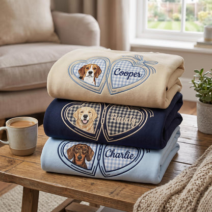 A stack of three folded sweatshirts in beige, navy blue, and light blue. Each features an embroidered heart-shaped locket design with a dog portrait on the left and a name on the right: Cooper, Buddy, and Charlie.