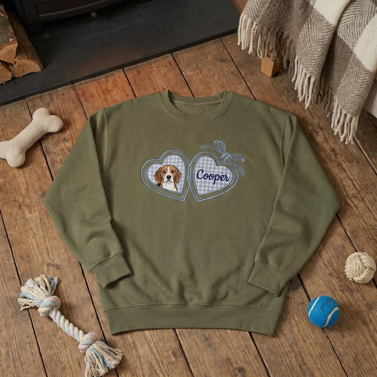 An olive green crewneck sweatshirt featuring a central graphic of an open heart-shaped locket. The left side of the locket displays a beagle dog, while the right side shows a blue and white gingham pattern with the name Cooper.