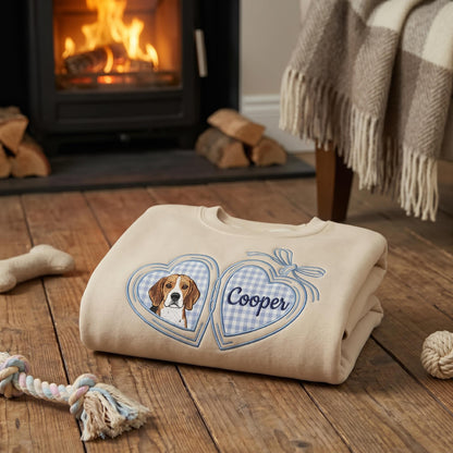 A beige crewneck sweatshirt featuring an embroidered design of two connected hearts. The left heart contains a portrait of a beagle, and the right heart displays the name Cooper in blue script, both set against a light blue gingham background.