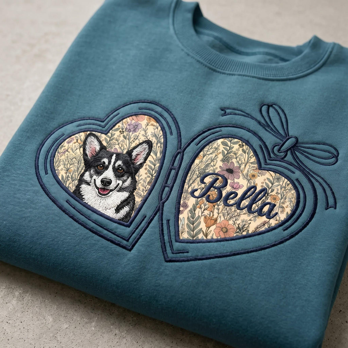 A teal crewneck sweatshirt featuring an embroidered design of an open heart-shaped locket. The left side displays a black and white Corgi, while the right side shows the name Bella in cursive script over a floral background.