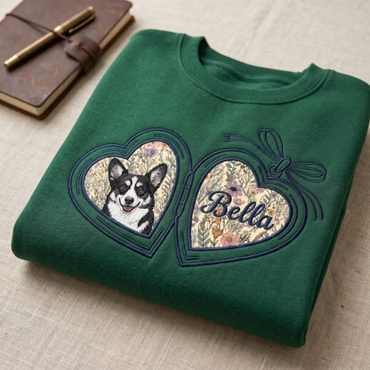 A dark green crewneck sweatshirt featuring an embroidered heart-shaped locket design. The left side displays a black and white Corgi, while the right side shows the name Bella in cursive script, both set against a floral background.