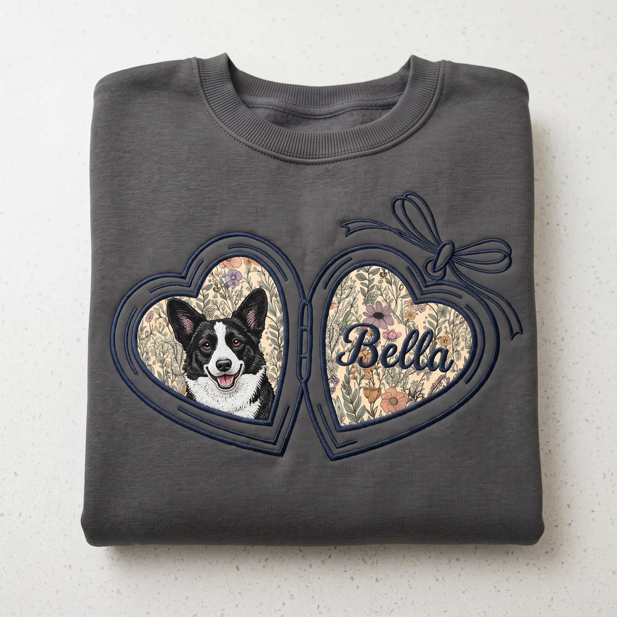 A charcoal gray crewneck sweatshirt featuring an embroidered heart-shaped locket design. The locket displays a black and white dog portrait on one side and the name Bella in cursive script on the other, both set against a floral background.