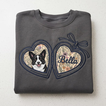 A charcoal gray crewneck sweatshirt featuring an embroidered heart-shaped locket design. The locket displays a black and white dog portrait on one side and the name Bella in cursive script on the other, both set against a floral background.
