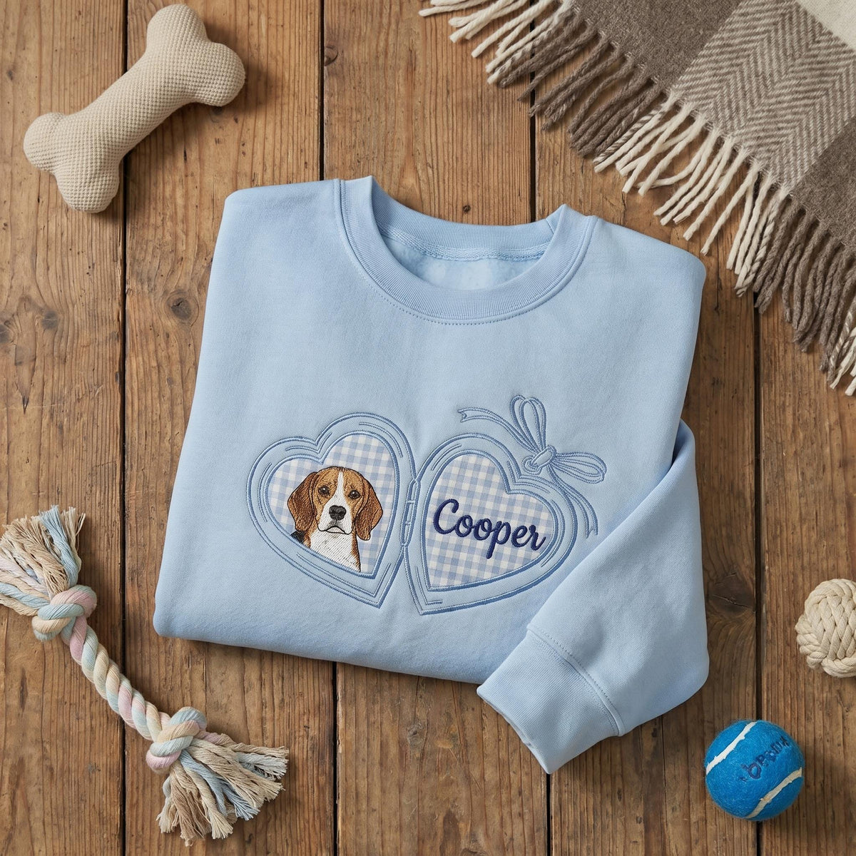 A light blue crewneck sweatshirt featuring an embroidered locket design. The left side of the locket displays a beagle dog, while the right side features a blue and white gingham pattern with the embroidered name Cooper.