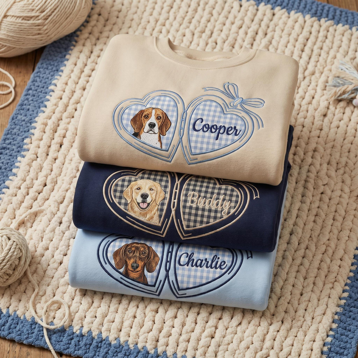 Three crewneck sweatshirts in beige, navy blue, and light blue, each featuring an embroidered locket design. The lockets contain illustrations of a beagle, a golden retriever, and a dachshund, with the names Cooper, Buddy, and Charlie.