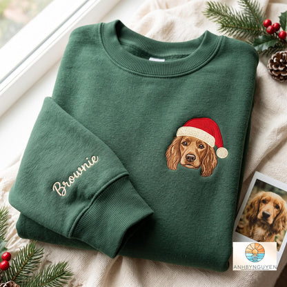 A dark green crewneck sweatshirt featuring an embroidered brown spaniel dog wearing a red Santa hat on the chest and the name Brownie embroidered in a cursive font on the sleeve cuff, showcasing a detailed canine portrait design.