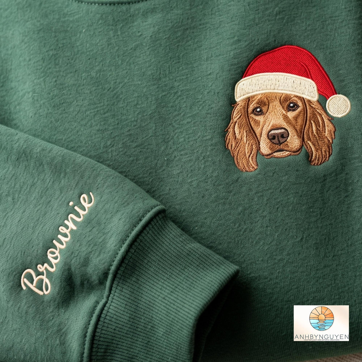 A dark green sweatshirt featuring an embroidered brown spaniel dog wearing a red and white Santa hat. The sleeve is embroidered with the name Brownie in a cursive font. A small logo with the text ANHBYNGUYEN is in the corner.