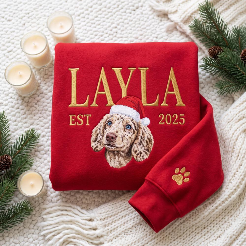 A red sweatshirt featuring gold embroidered text that reads LAYLA EST 2025. Centered is a detailed illustration of a spotted puppy wearing a red and white Santa hat. A small gold embroidered paw print is visible on the sleeve cuff.