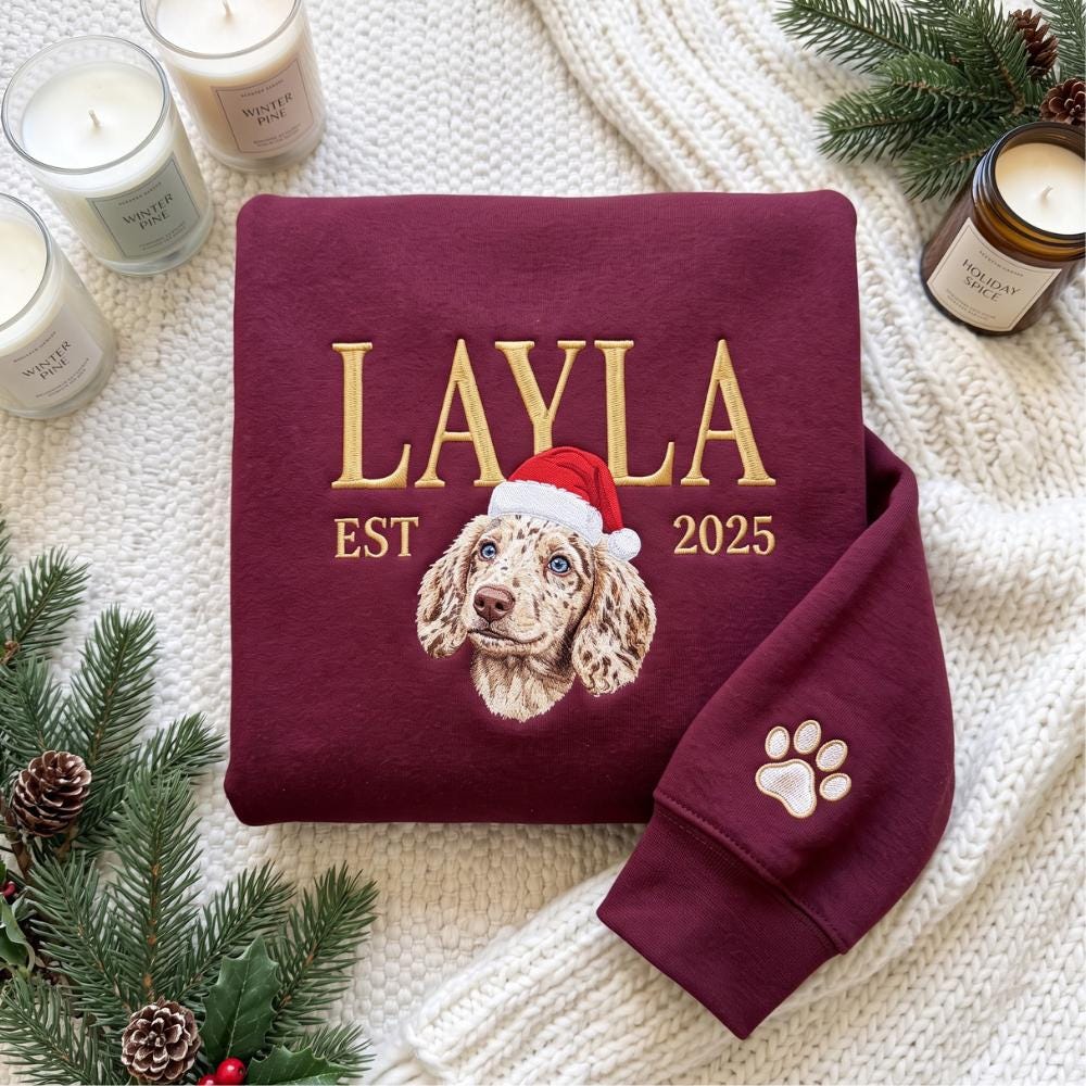 A maroon sweatshirt featuring gold embroidered text that reads LAYLA EST 2025. Centered is an embroidered portrait of a speckled dog wearing a red and white Santa hat. A white embroidered paw print is visible on the sleeve cuff.
