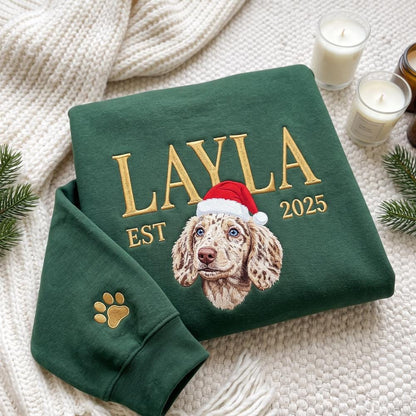 A dark green sweatshirt featuring gold embroidered text that reads LAYLA EST 2025. Centered is an embroidered portrait of a spotted dog wearing a red and white Santa hat. A gold embroidered paw print is on the sleeve cuff.