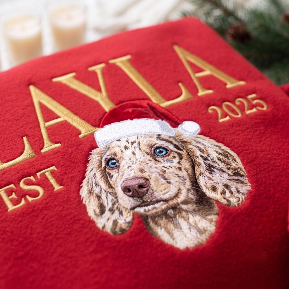 A red fleece blanket featuring an embroidered portrait of a speckled dog with blue eyes wearing a red and white Santa hat. The text LAYLA and 2025 are embroidered in gold thread above the dog, with the letters EST visible below.