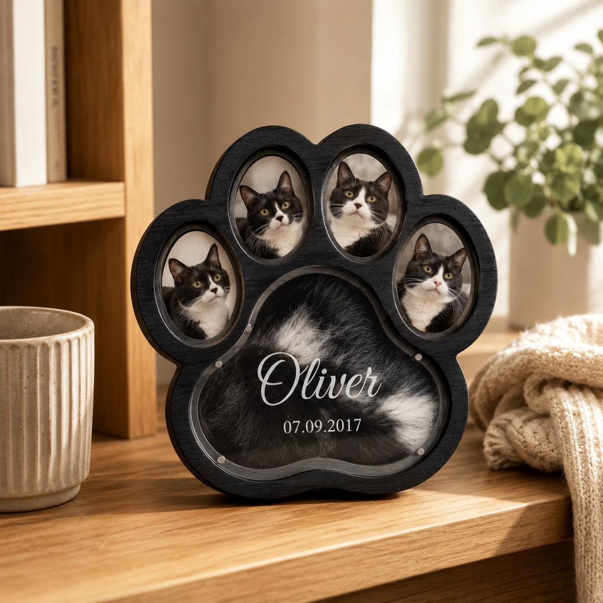 A black wooden paw-shaped picture frame featuring four oval photo slots containing images of a black and white cat. The central area displays the name Oliver and the date 07.09.2017 in white script and sans-serif lettering.