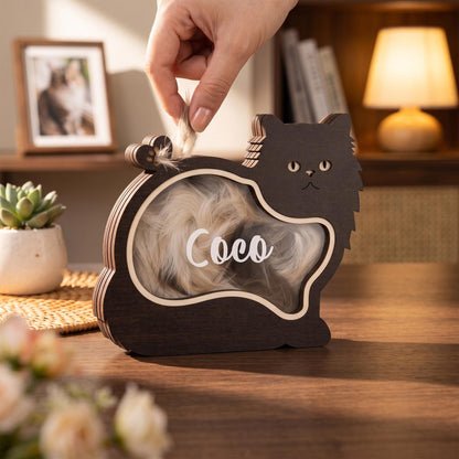 A dark brown wooden cat-shaped shadow box with a clear front panel featuring the name Coco in white script. The interior contains light-colored fur, and the frame has a layered wood construction with a cutout cat silhouette design.