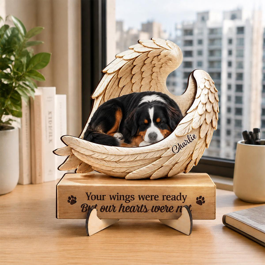 A wooden memorial sculpture featuring a Bernese Mountain dog resting inside a pair of carved angel wings. The base is engraved with the text "Your wings were ready But our hearts were not" and the name "Charlie" is on the wing.