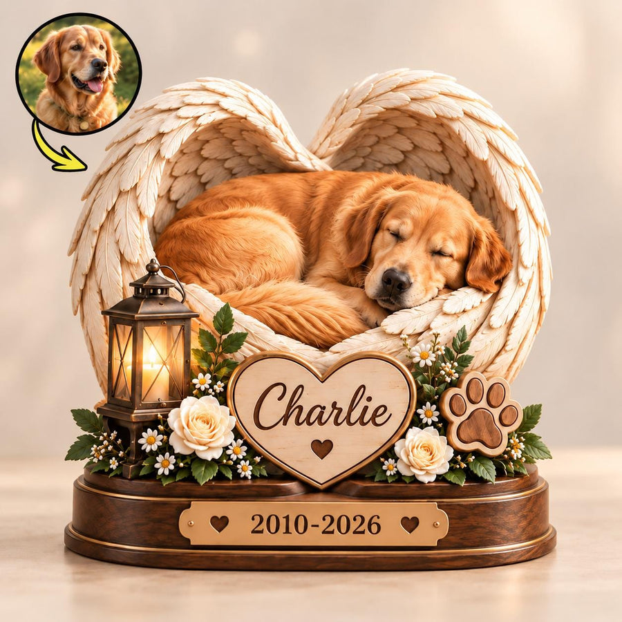A memorial sculpture featuring a golden retriever sleeping inside a pair of large, cream-colored angel wings. A heart-shaped plaque displays the name Charlie, accompanied by a lantern, white roses, and a paw print. A base shows 2010-2026.