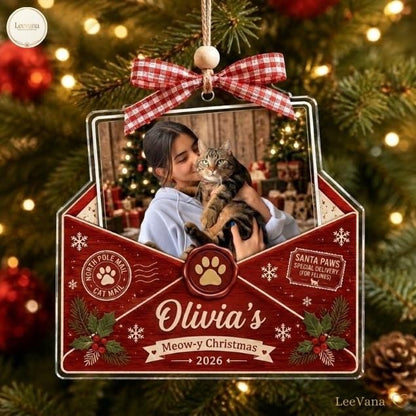 A custom envelope-shaped Christmas ornament featuring a photo of a person holding a tabby cat. The red design includes a red and white gingham bow, a gold paw print seal, and text reading Olivia's Meow-y Christmas 2026, North Pole Mail, and Santa Paws.