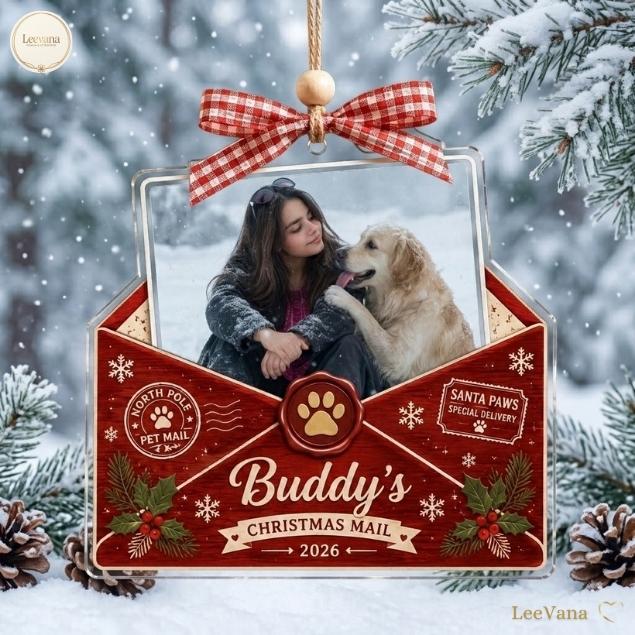 A clear acrylic envelope-shaped Christmas ornament featuring a red wood-grain pattern, a red and white gingham bow, and a photo of a person with a golden retriever. Text includes North Pole Pet Mail, Santa Paws, and Buddy's Christmas Mail 2026.
