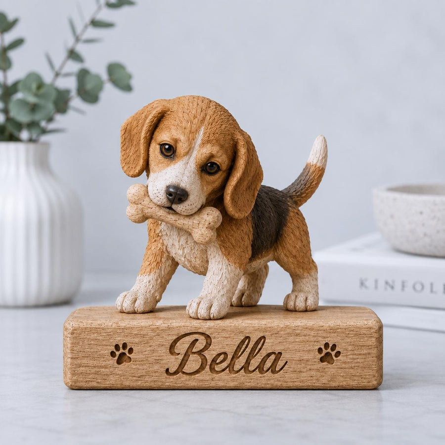A beagle puppy figurine with brown, white, and black fur holding a bone in its mouth. The puppy stands on a rectangular wooden base engraved with the name Bella and two paw prints, one on each side of the name in a script font.