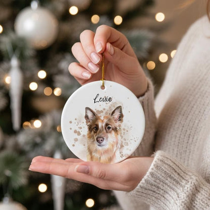 A round, white ceramic Christmas ornament featuring a watercolor portrait of a dog with brown and white fur. The dog's name, "Louie," is written above the portrait in black script. The ornament is held by a person.