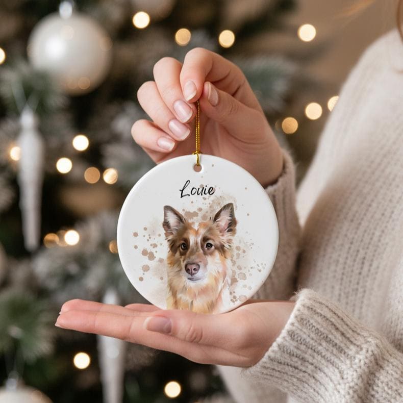 A round, white ceramic Christmas ornament featuring a watercolor portrait of a dog with brown and white fur. The dog's name, "Louie," is written above the portrait in black script. The ornament is held by a person.