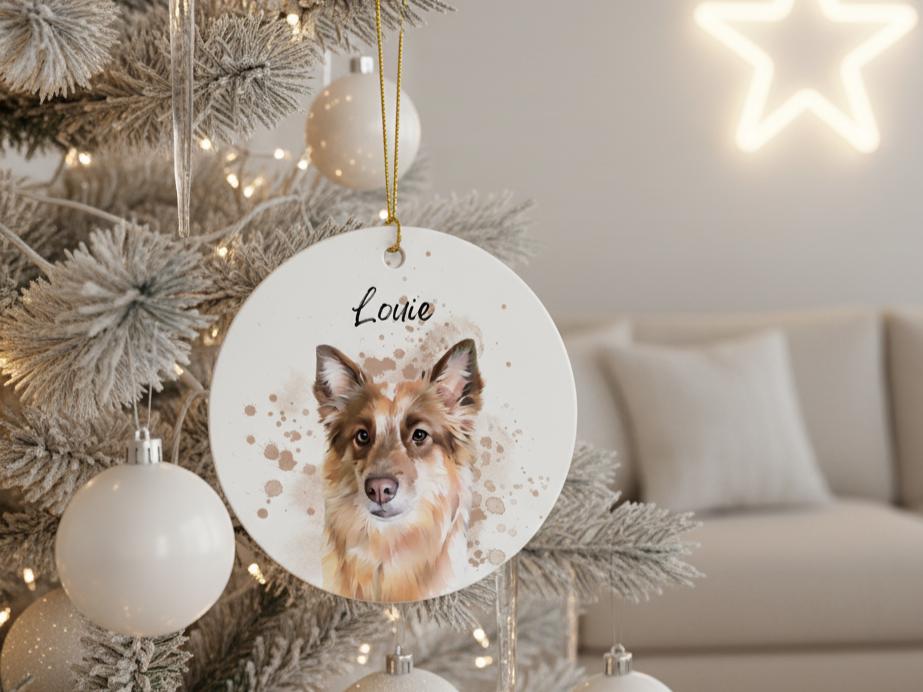 A round, white Christmas ornament featuring a watercolor portrait of a dog with brown and white fur. The dog's name, "Louie," is written above the portrait in black script. The ornament hangs from a gold string.