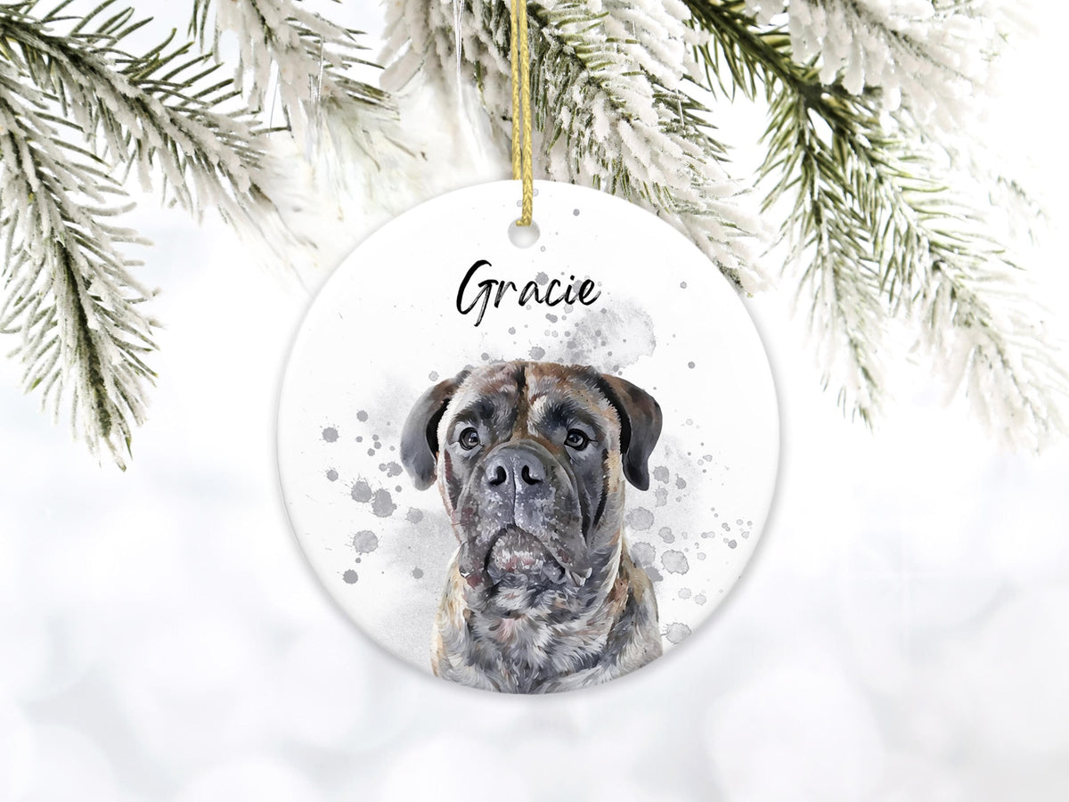 A round, white ceramic ornament featuring a watercolor portrait of a brindle dog with the name "Gracie" in black script. The ornament has a gold string for hanging and is set against a snowy background.