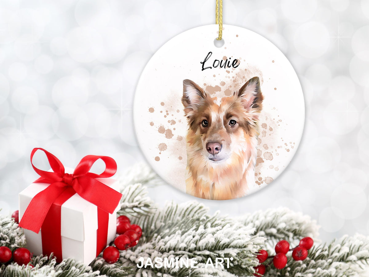 A round, white Christmas ornament featuring a watercolor portrait of a dog with brown and white fur, with the name "Louie" in script. A gift box with a red ribbon and faux evergreen branches with red berries are in the foreground.
