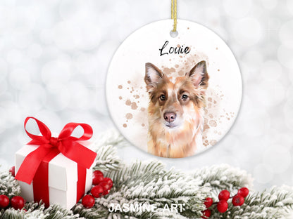 A round, white Christmas ornament featuring a watercolor portrait of a dog with brown and white fur, with the name "Louie" in script. A gift box with a red ribbon and faux evergreen branches with red berries are in the foreground.