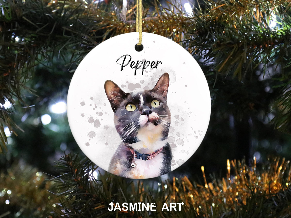 A round, white ceramic Christmas ornament featuring a watercolor portrait of a cat named Pepper. The cat has black, white, and gray fur, with green eyes. The ornament is hanging from a gold string and has the text "Pepper" and "JASMINE ART".