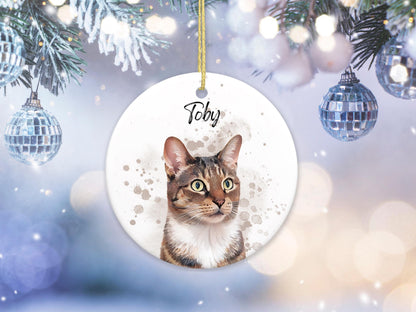 A round, white Christmas ornament featuring a watercolor portrait of a tabby cat with the name "Toby" in script. The cat has green eyes and brown and white fur. The ornament hangs from a gold string.