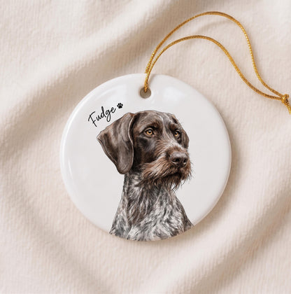 A round white ceramic ornament featuring a detailed illustration of a brown and white German Wirehaired Pointer dog. The name Fudge is printed in black script next to a small paw print icon. A gold string is attached for hanging.
