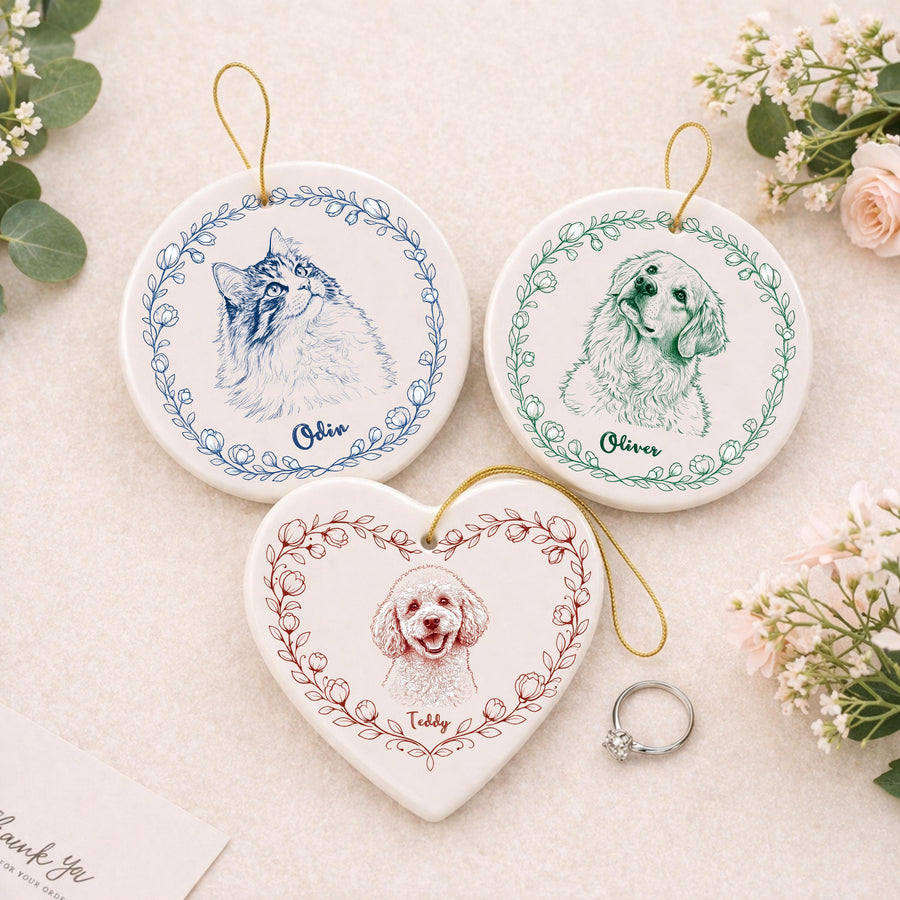 Two white ceramic circular ornaments and one white ceramic heart-shaped ornament, each featuring a detailed pet portrait sketch surrounded by a floral wreath and a gold hanging cord. The names Odin, Oliver, and Teddy are printed below.