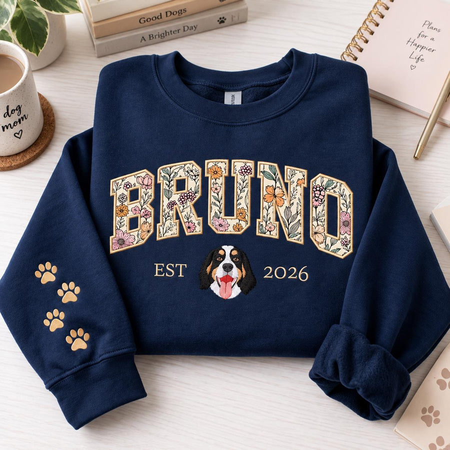 A navy blue crewneck sweatshirt featuring the name BRUNO in large block letters filled with a floral pattern. Below the name is a graphic of a dog face with the text EST 2026. The left sleeve displays four small gold paw prints.