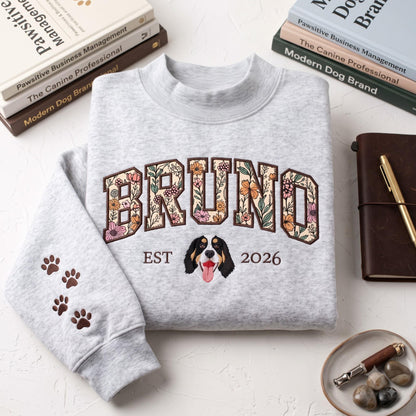 A light gray crewneck sweatshirt featuring the name BRUNO in large block letters filled with a colorful floral pattern. Below the name is an illustration of a dog face, the text EST 2026, and brown paw prints on the left sleeve.
