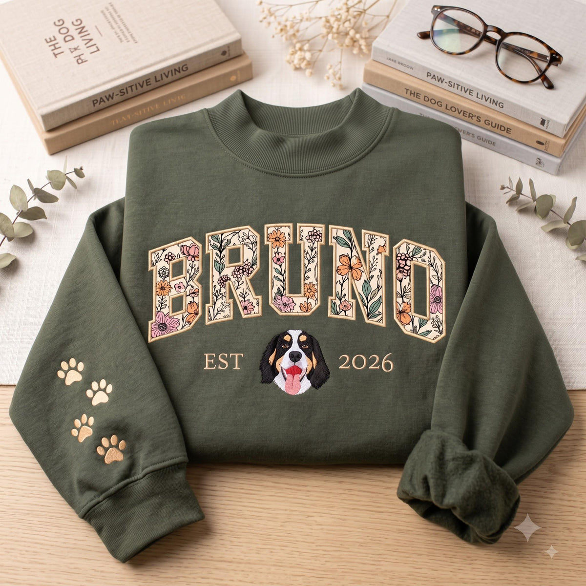 An olive green crewneck sweatshirt featuring the name BRUNO in large, floral-patterned block letters across the chest. Below the name is the text EST 2026 with a dog face illustration in the center. The left sleeve has gold paw prints.