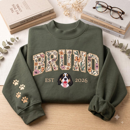 An olive green crewneck sweatshirt featuring the name BRUNO in large, floral-patterned block letters across the chest. Below the name is the text EST 2026 with a dog face illustration in the center. The left sleeve has gold paw prints.