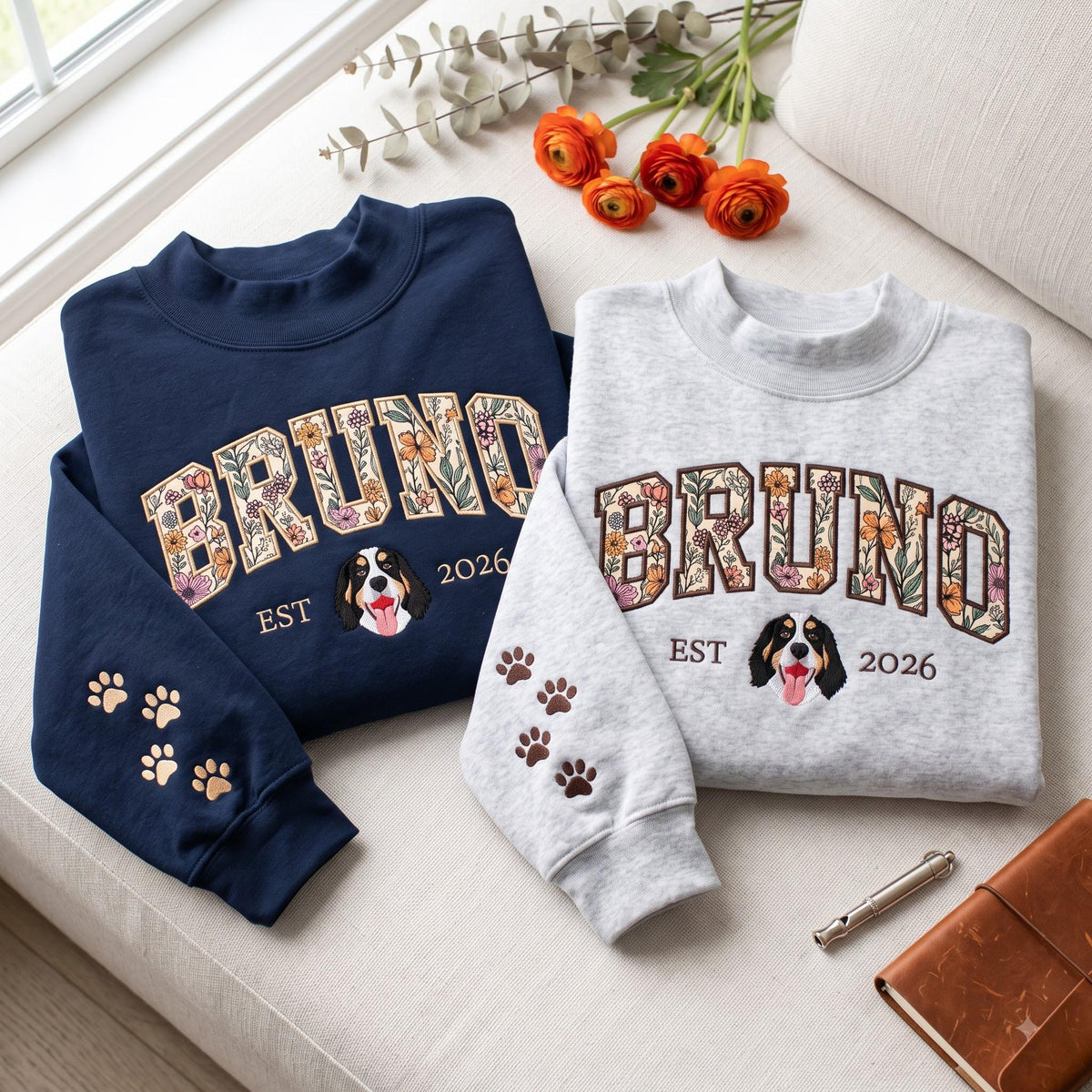 A navy blue and a light gray sweatshirt, each featuring the name BRUNO in floral-patterned block letters. Below the name is an embroidered Bernese Mountain Dog face, the text EST 2026, and paw prints on the sleeve cuffs.