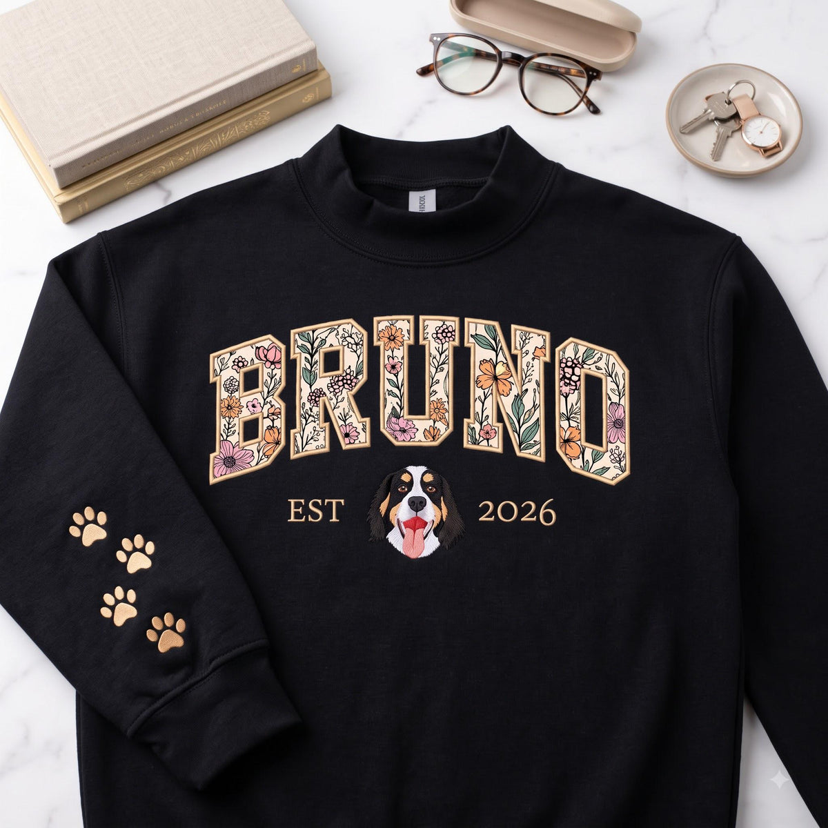 A black long-sleeve sweatshirt featuring the name BRUNO in large block letters filled with a floral pattern. Below the name is a graphic of a dog face, the text EST 2026, and four gold-colored paw prints on the left sleeve.