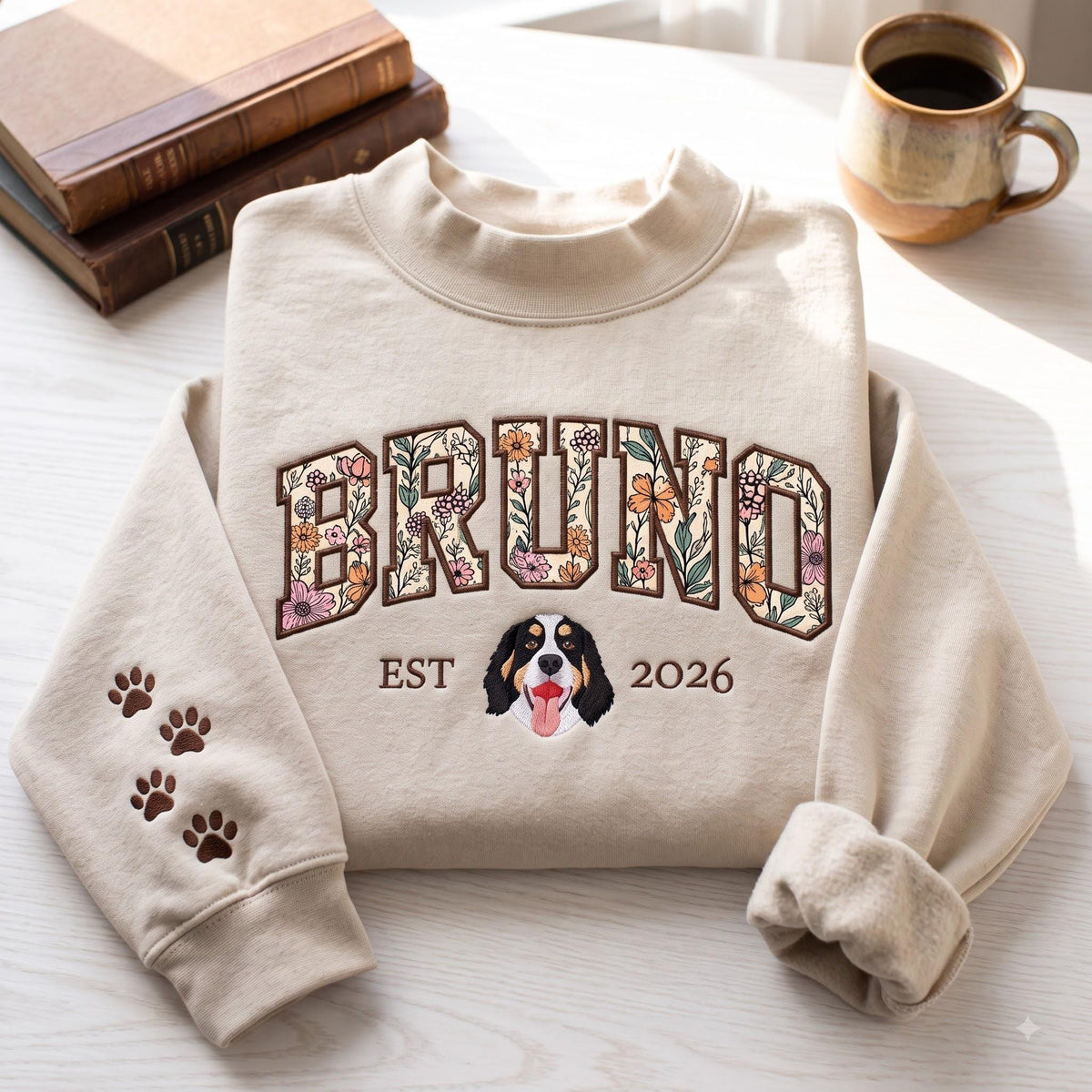 A beige crewneck sweatshirt featuring the name BRUNO in large block letters filled with a colorful floral pattern. Below the name is the text EST 2026 flanking an embroidered Bernese Mountain Dog face. The left sleeve has paw prints.