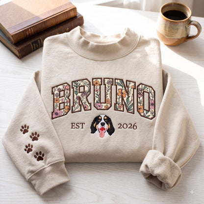 A beige crewneck sweatshirt featuring the name BRUNO in large block letters filled with a colorful floral pattern. Below the name is the text EST 2026 flanking an embroidered Bernese Mountain Dog face. The left sleeve has paw prints.