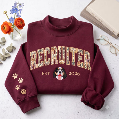A maroon mock neck sweatshirt featuring the word RECRUITER in a floral-patterned font across the chest. Below the text is a graphic of a Bernese Mountain Dog face flanked by the text EST 2026. The left sleeve has three gold paw prints.