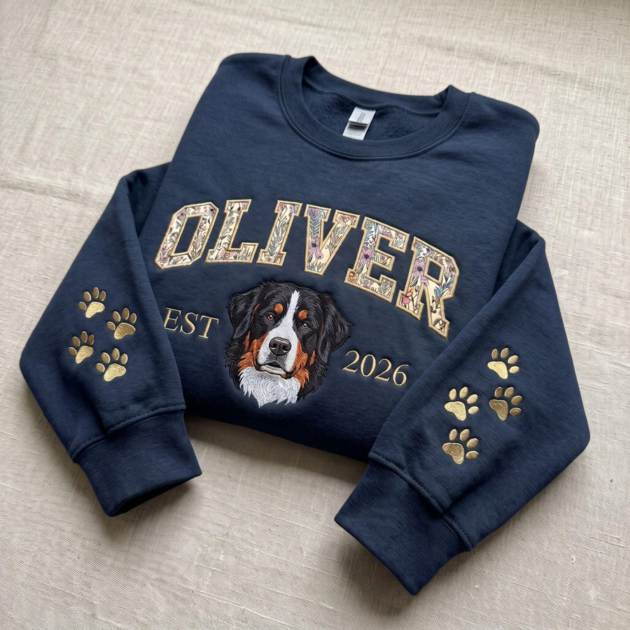 A navy blue crewneck sweatshirt featuring the name OLIVER in floral-patterned block letters across the chest. Below the name is a portrait of a Bernese Mountain Dog, the text EST 2026, and gold embroidered paw prints on both sleeves.