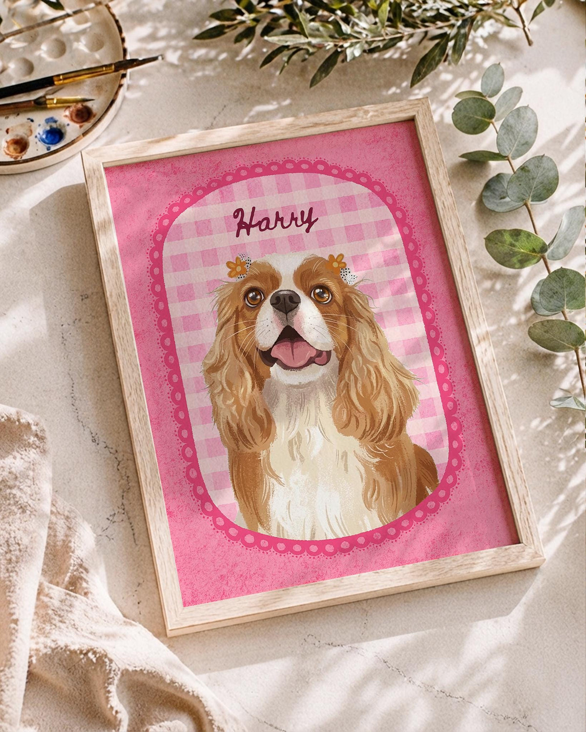 A framed print featuring a digital illustration of a brown and white Cavalier King Charles Spaniel with small yellow flowers on its head. The dog is centered within a pink gingham oval frame with the name Harry written above it.