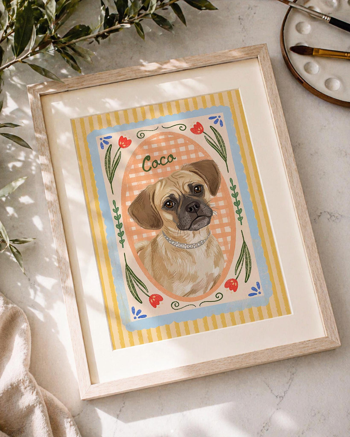 A framed art print featuring a tan dog with a dark muzzle wearing a silver collar, centered in an oval frame against a peach gingham background. The border includes yellow stripes, blue accents, and red tulip illustrations with the name Coco.