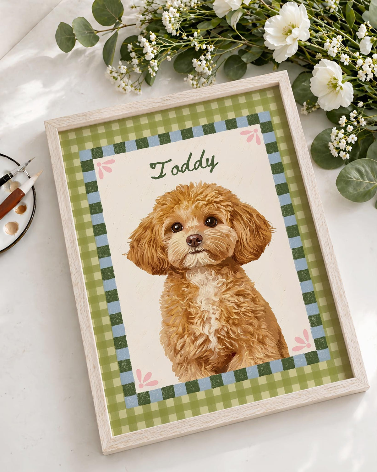 A framed portrait of a fluffy golden-brown dog with a white chest patch. The illustration features the name Toddy in green script, set against a cream background with a green and blue checkered border and small pink flower accents.