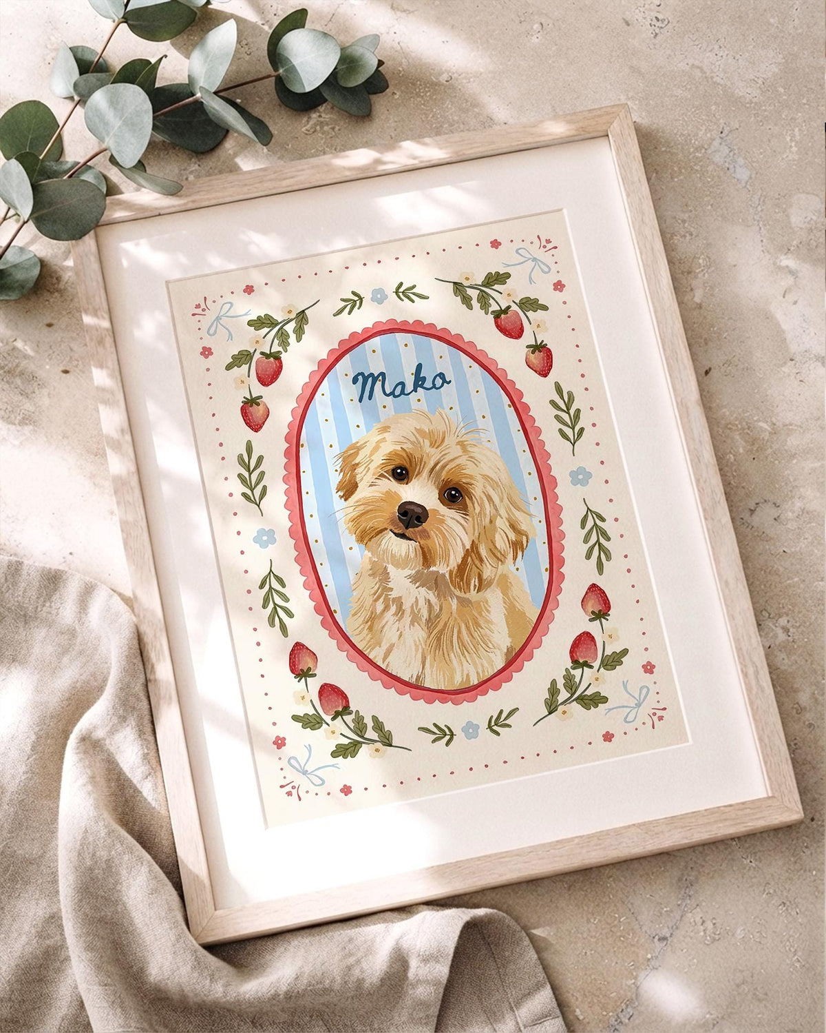 A framed illustration of a light brown dog inside a blue and white striped oval frame with a red scalloped border. The name Mako is written in blue script above the dog. The border features red strawberries, green leaves, and small blue flowers.