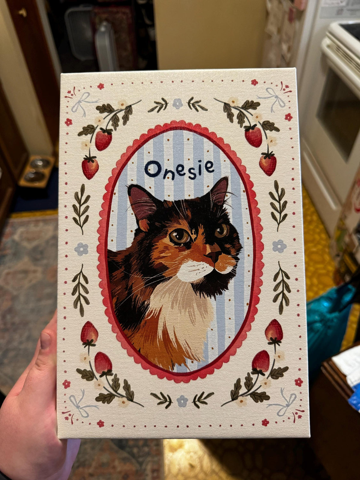 A rectangular canvas print featuring a portrait of a calico cat inside a red scalloped oval frame against a light blue striped background. The word Onesie is printed above the cat. The border is decorated with strawberries and bows.